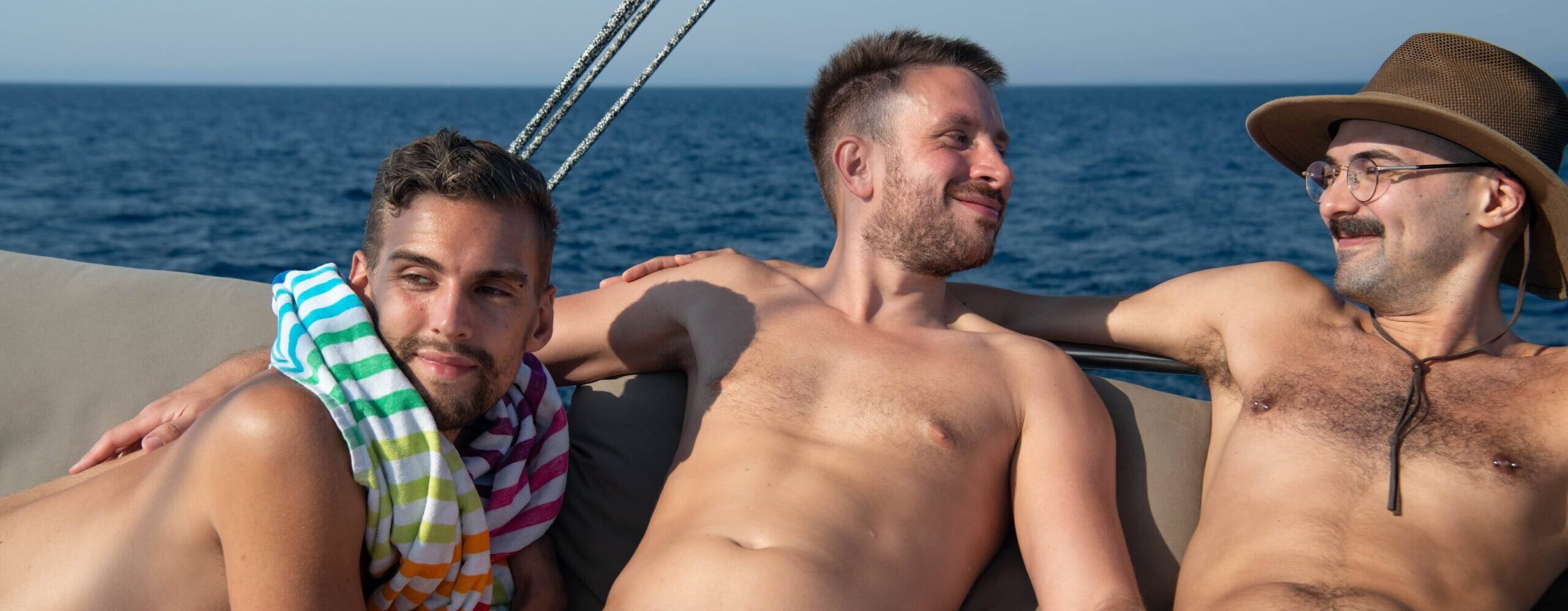 Three gay naturist Saltyboys lounging on catamaran daybeds